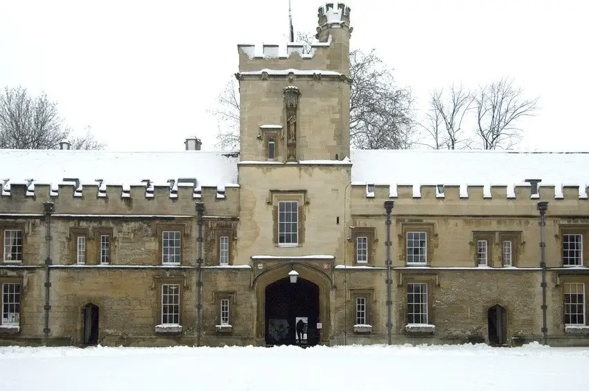 Front Quad at St John's College under snow: the gate tower with its carved statue, battlemented ranges either side, and the lawn covered white.